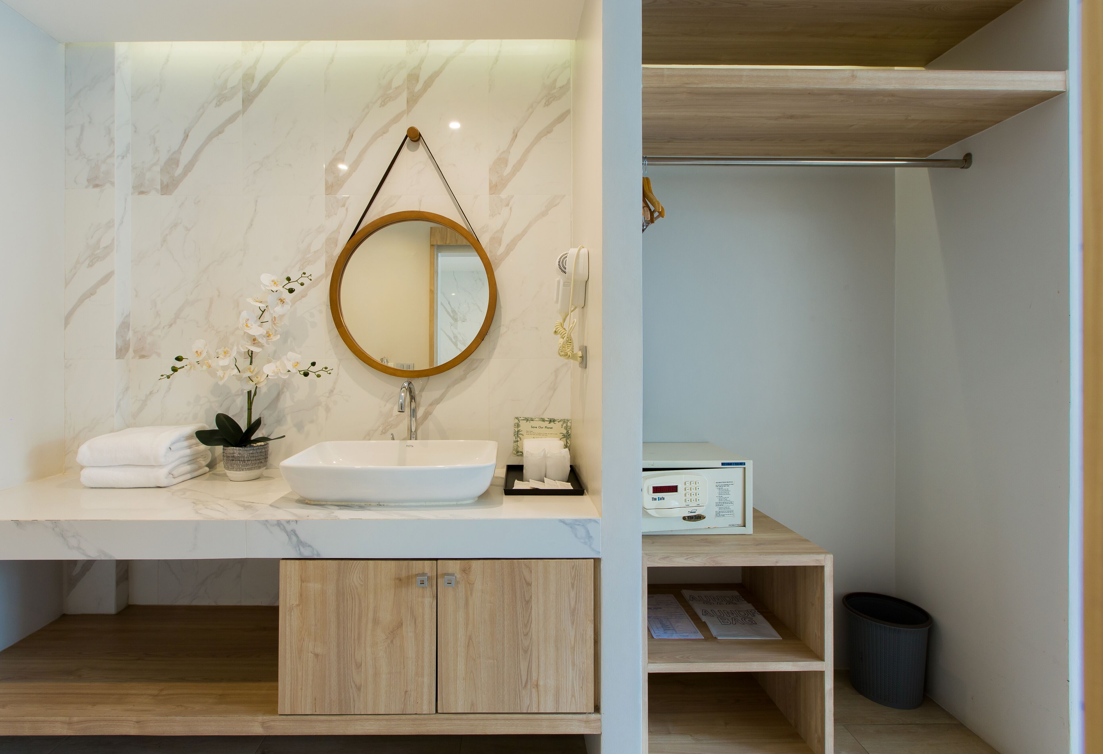 A bathroom with a marble vanity, a round mirror, a white sink, a towel rack, a small potted plant, and a waste bin.