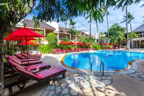 A swimming pool with red umbrellas and lounge chairs, surrounded by palm trees and a building with a tiled roof. - Coconut Village Resort (Patong)