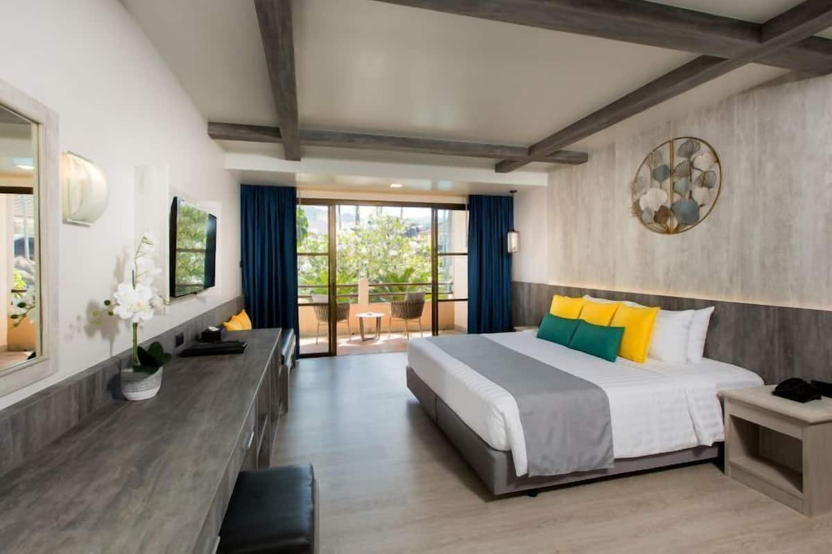 Una habitación de hotel moderna con una cama grande, un escritorio con silla, una mesita pequeña y un balcón con mobiliario exterior.