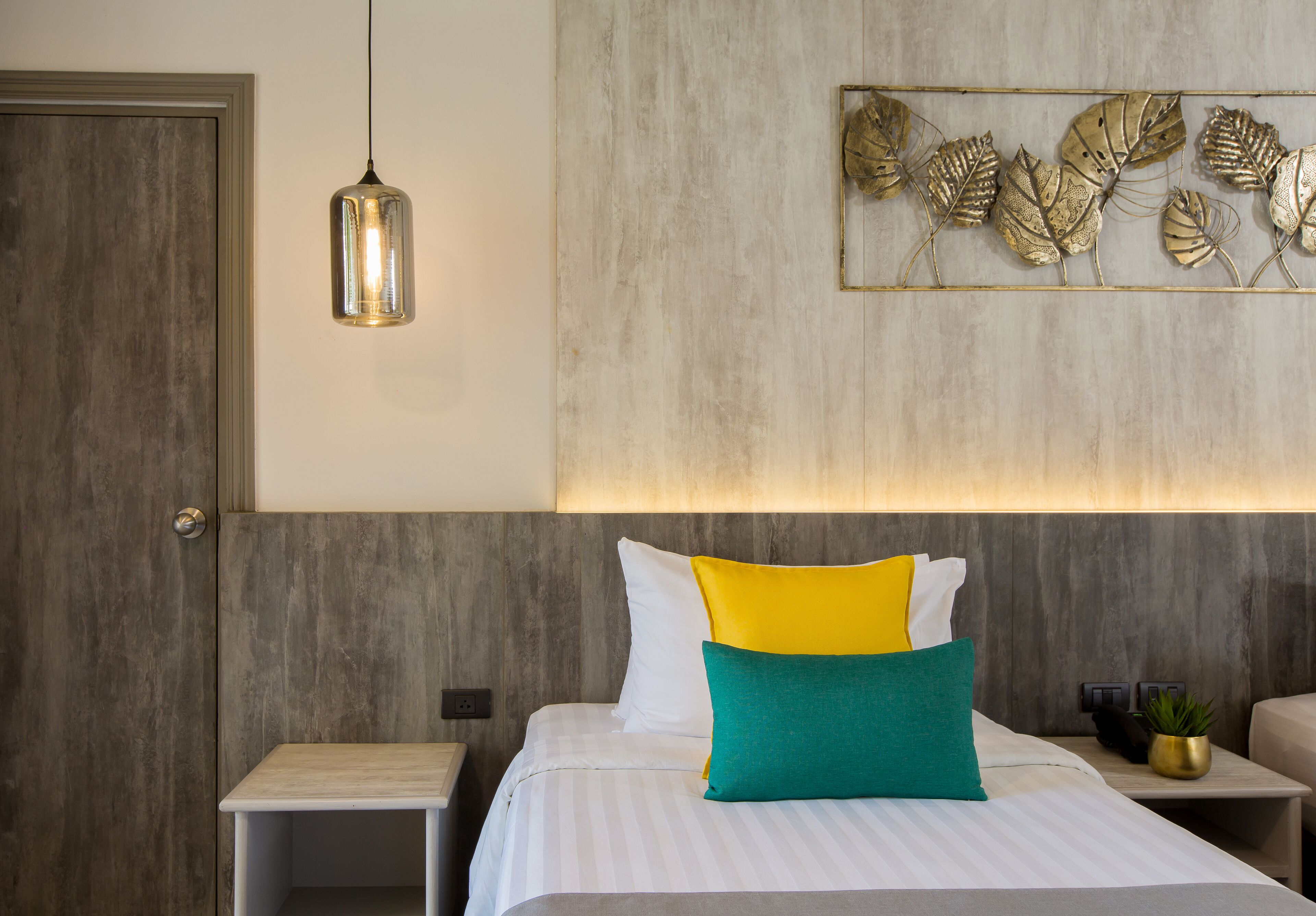 Una habitación de hotel con cama, mesitas de noche, lámpara de pared y una obra de arte decorativa con hojas.