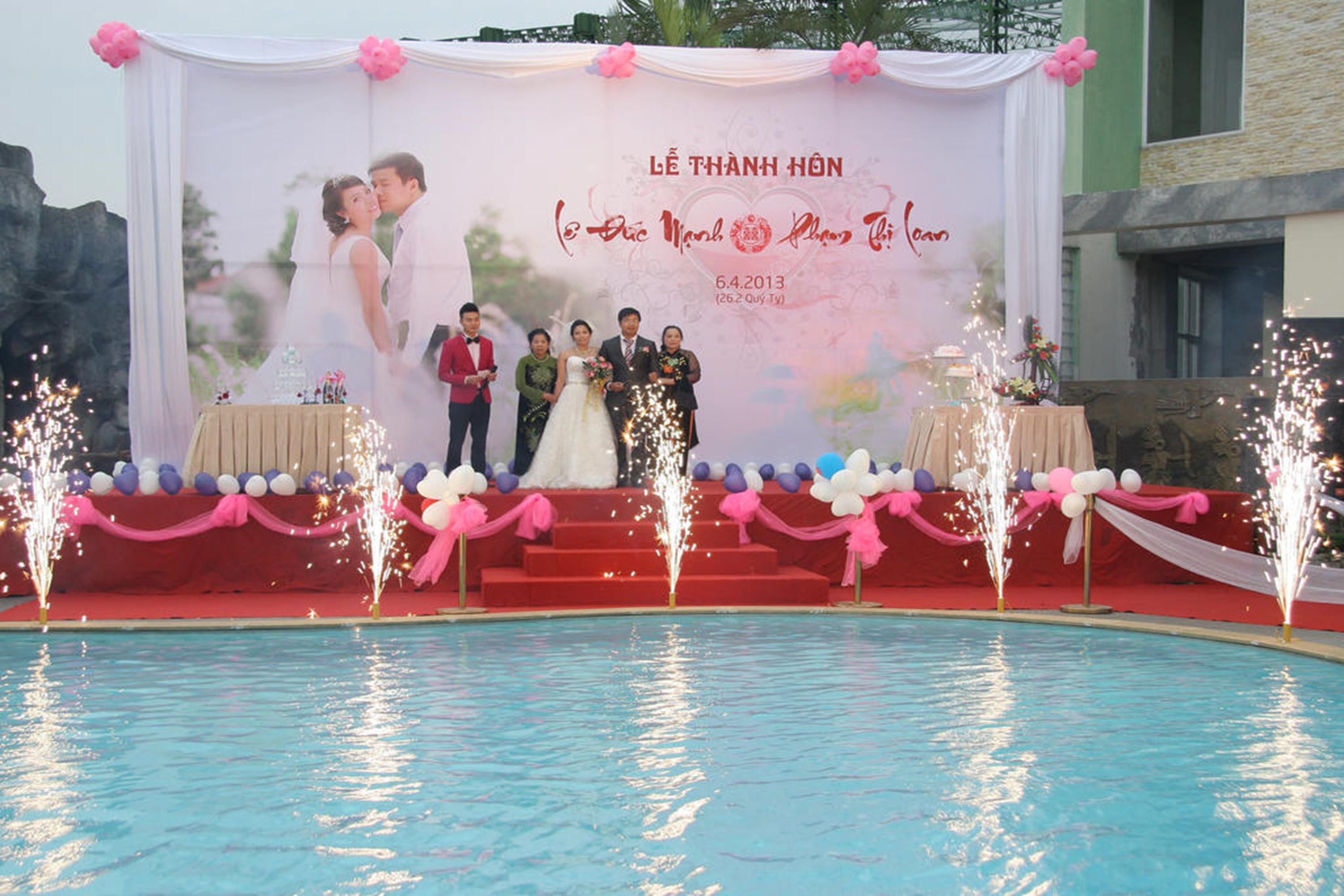 A wedding ceremony with a backdrop featuring a couple and guests, a pool with water fountains, and a stage with a table and chairs.