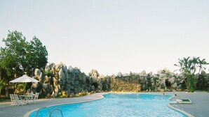 A swimming pool with a curved edge, surrounded by a rocky wall and palm trees. There are lounge chairs and an umbrella near the pool. - Camela Hotel & Resort (Hai Phong)