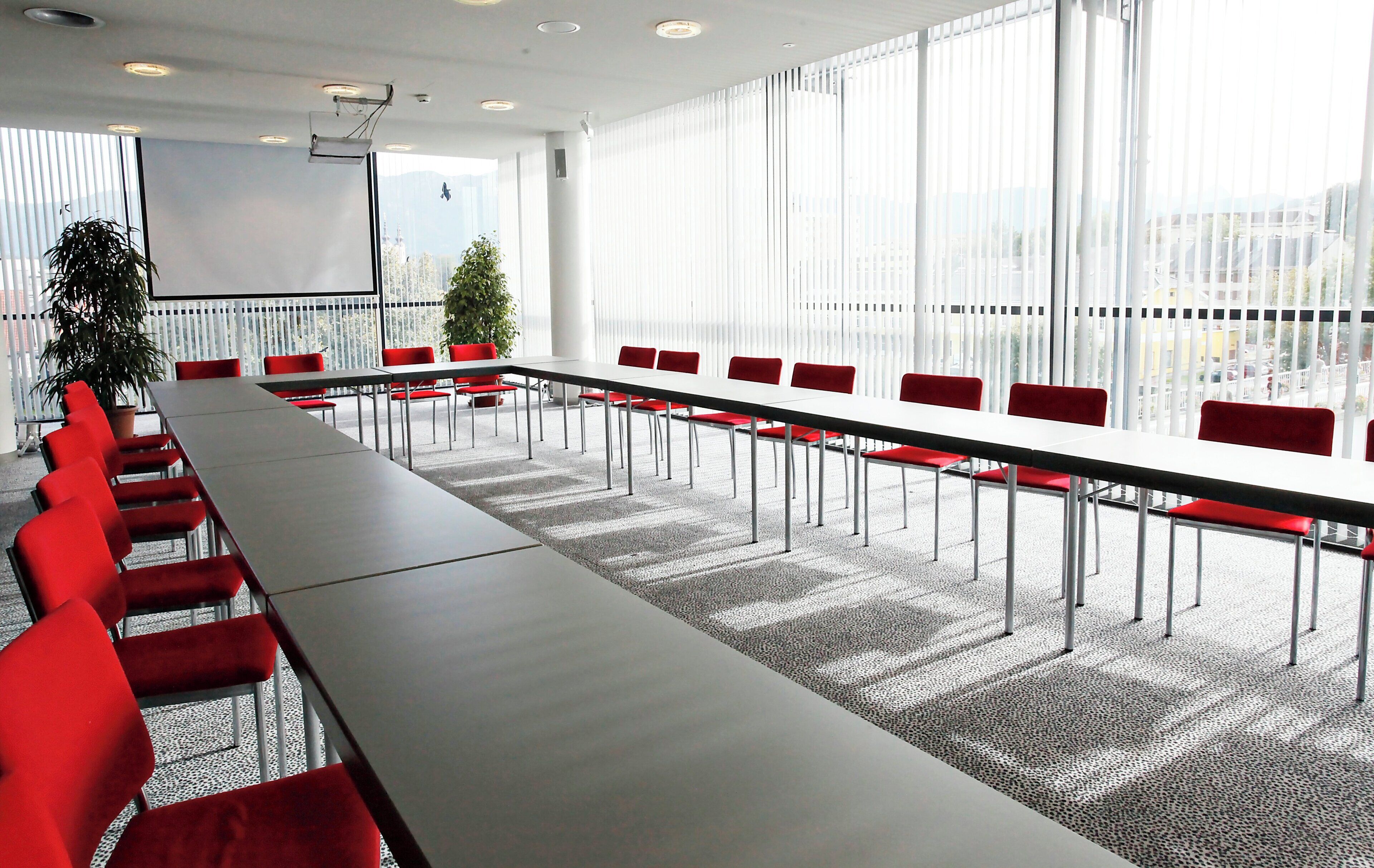 Une salle de conférence avec une longue table, des chaises rouges, un écran de projection et de grandes fenêtres.