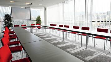 Une salle de conférence avec une longue table, des chaises rouges, un écran de projection et de grandes fenêtres.