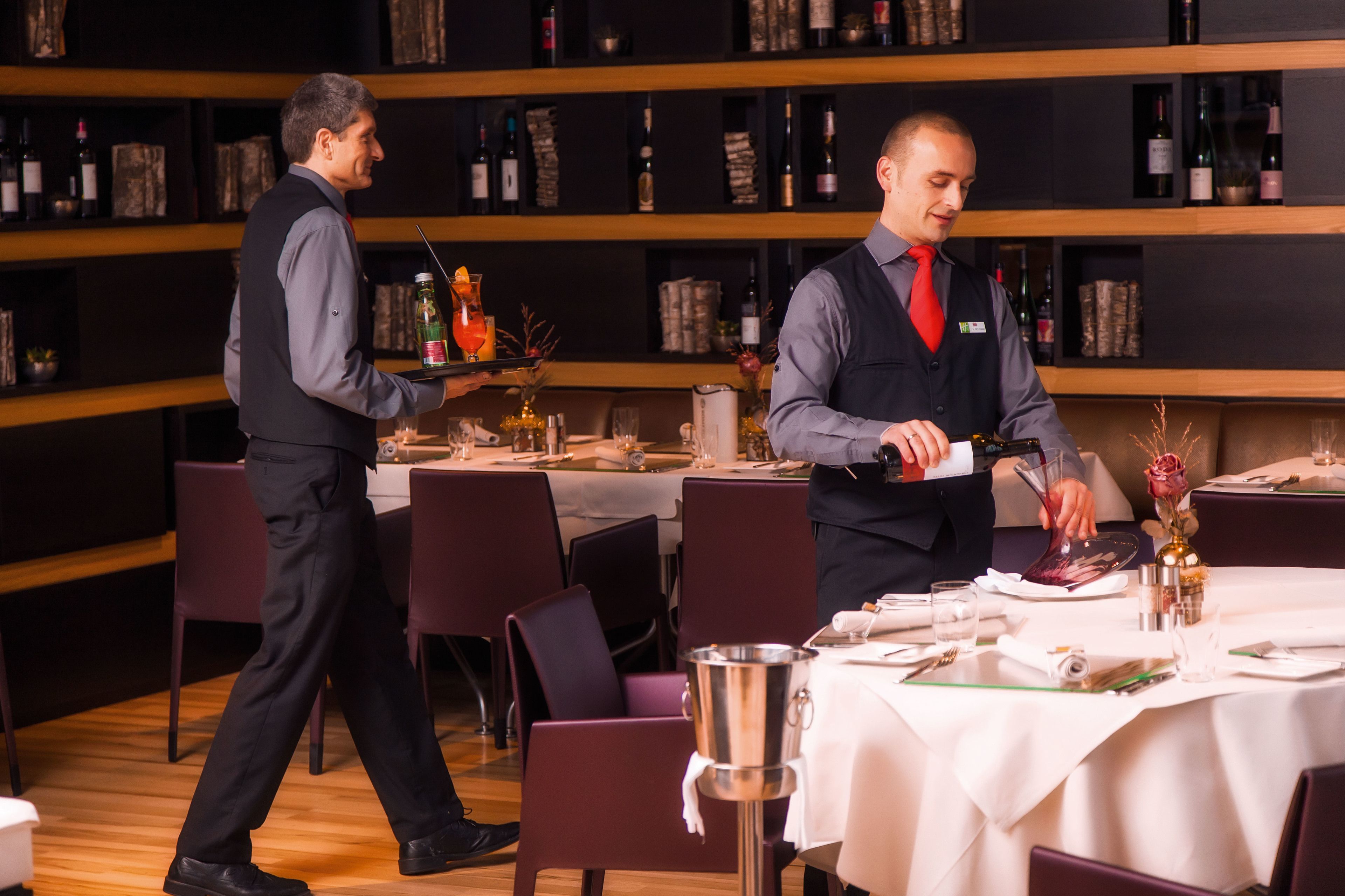A restaurant with a wooden floor, tables set with white tablecloths, and a bar area with shelves of bottles.