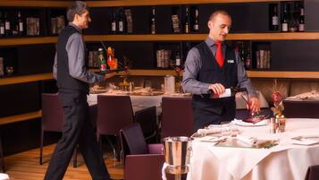 A restaurant with a wooden floor, tables set with white tablecloths, and a bar area with shelves of bottles.