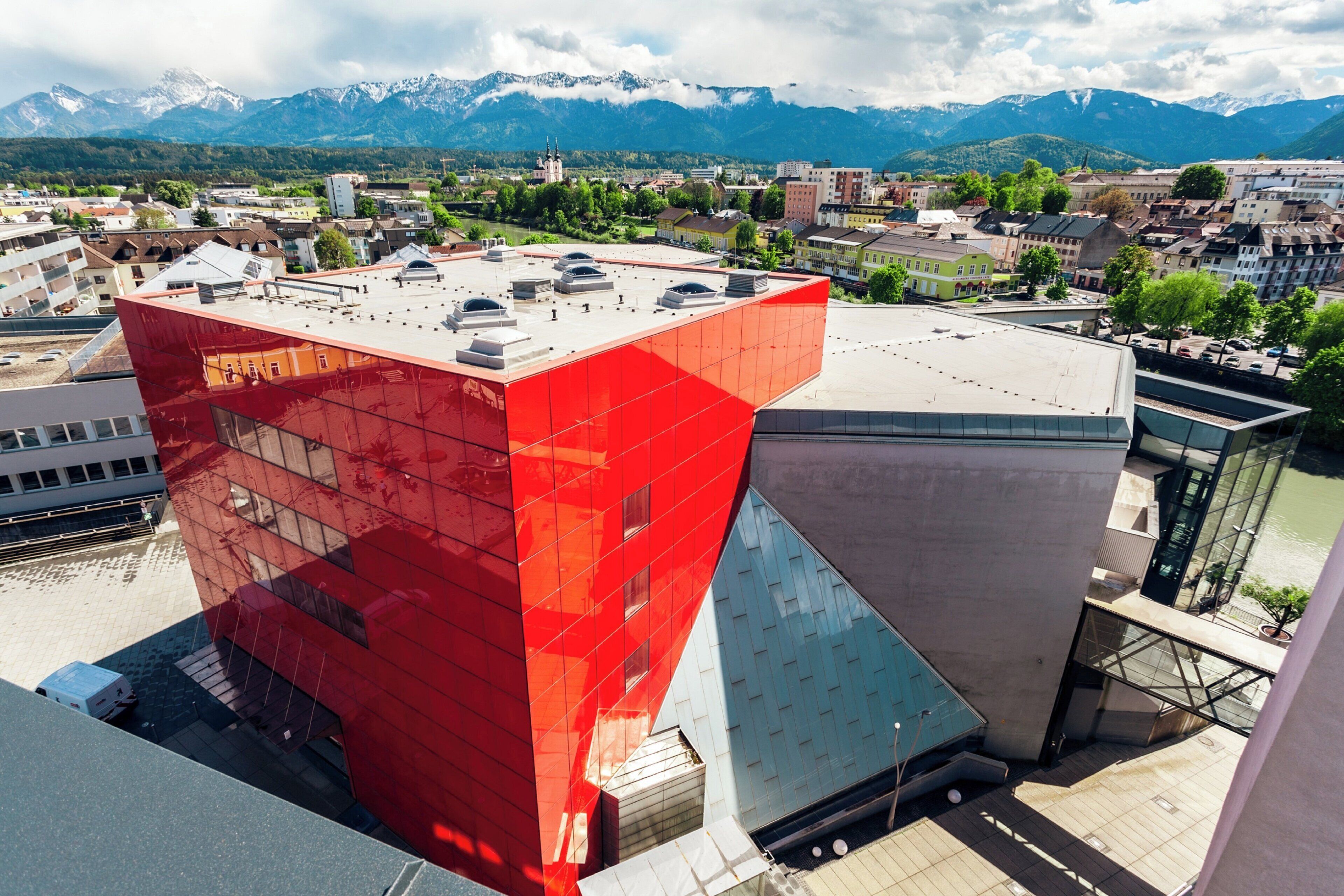 Un edificio moderno con fachada roja, rodeado de un paisaje urbano y montañas al fondo.