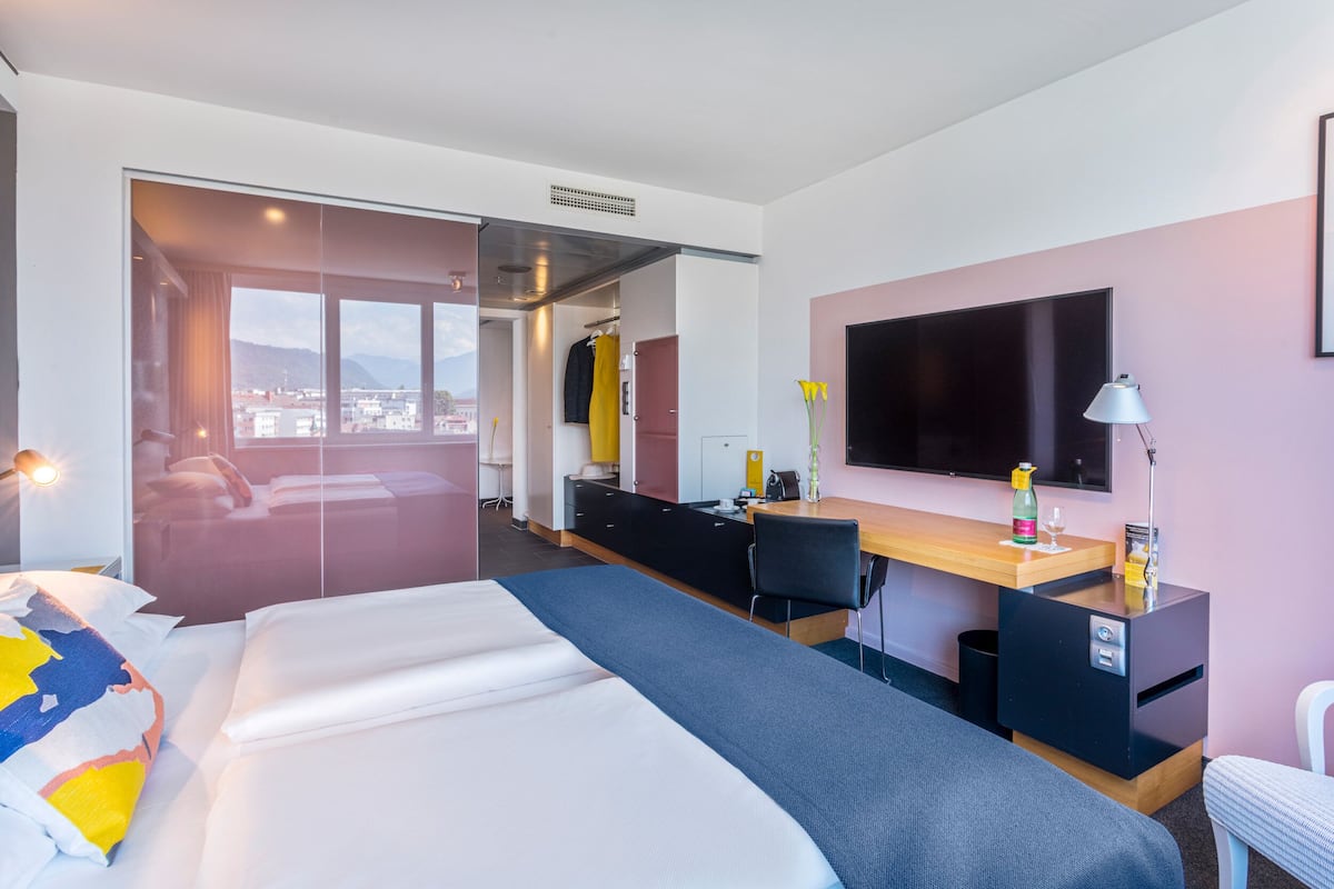 Ein Hotelzimmer mit einem großen Bett, einem Schreibtisch, einem Stuhl, einem Fernseher und Blick auf die Stadt.