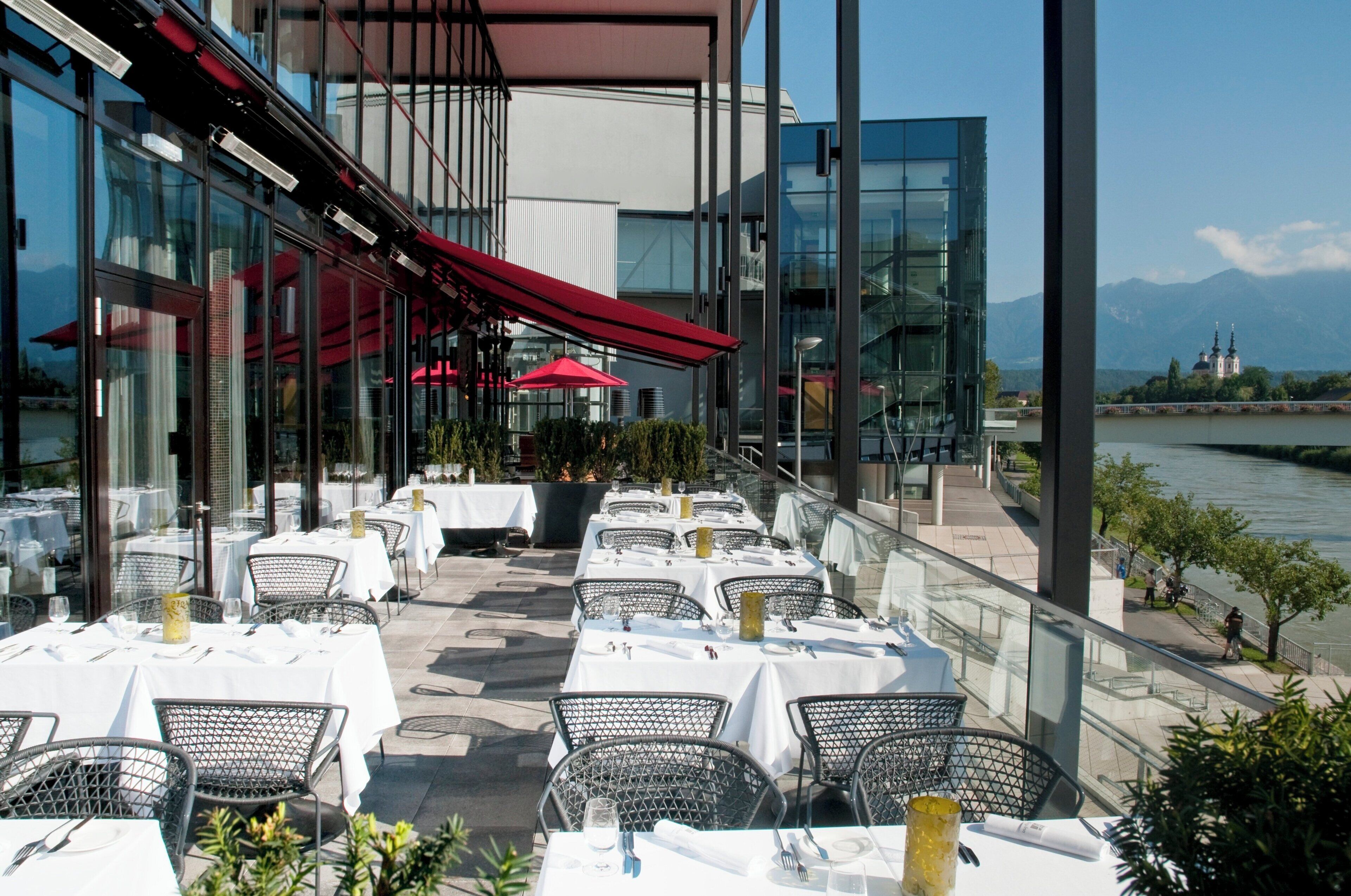 Un área de comedor al aire libre con mesas y sillas, vista al río y un edificio con ventanales amplios.