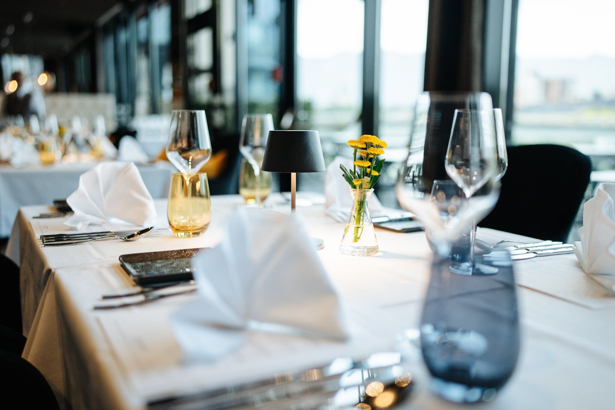 A dining table set with white tablecloth, wine glasses, a vase with yellow flowers, and a black lamp.