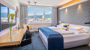 Habitación de hotel con una cama grande, un escritorio con silla, vistas a la ciudad y una ventana con cortinas.