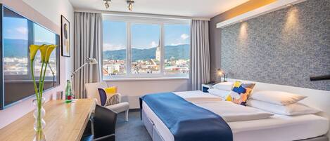 A hotel room with a large bed, a desk with a chair, a view of the city, and a window with curtains.