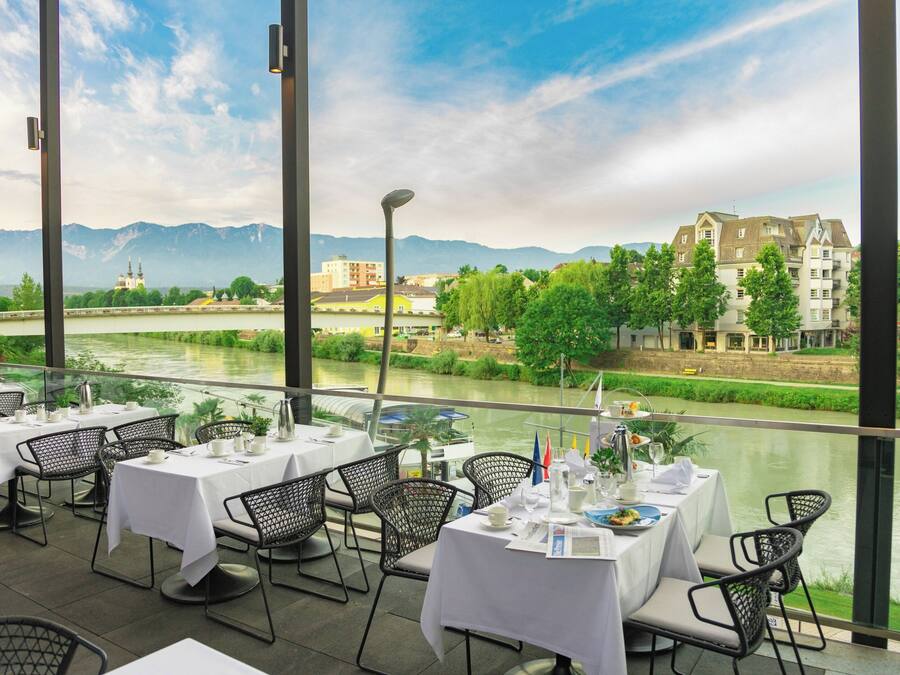 An outdoor dining area with tables and chairs, overlooking a river and buildings in the background.