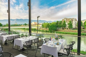 An outdoor dining area with tables and chairs, overlooking a river and buildings in the background.