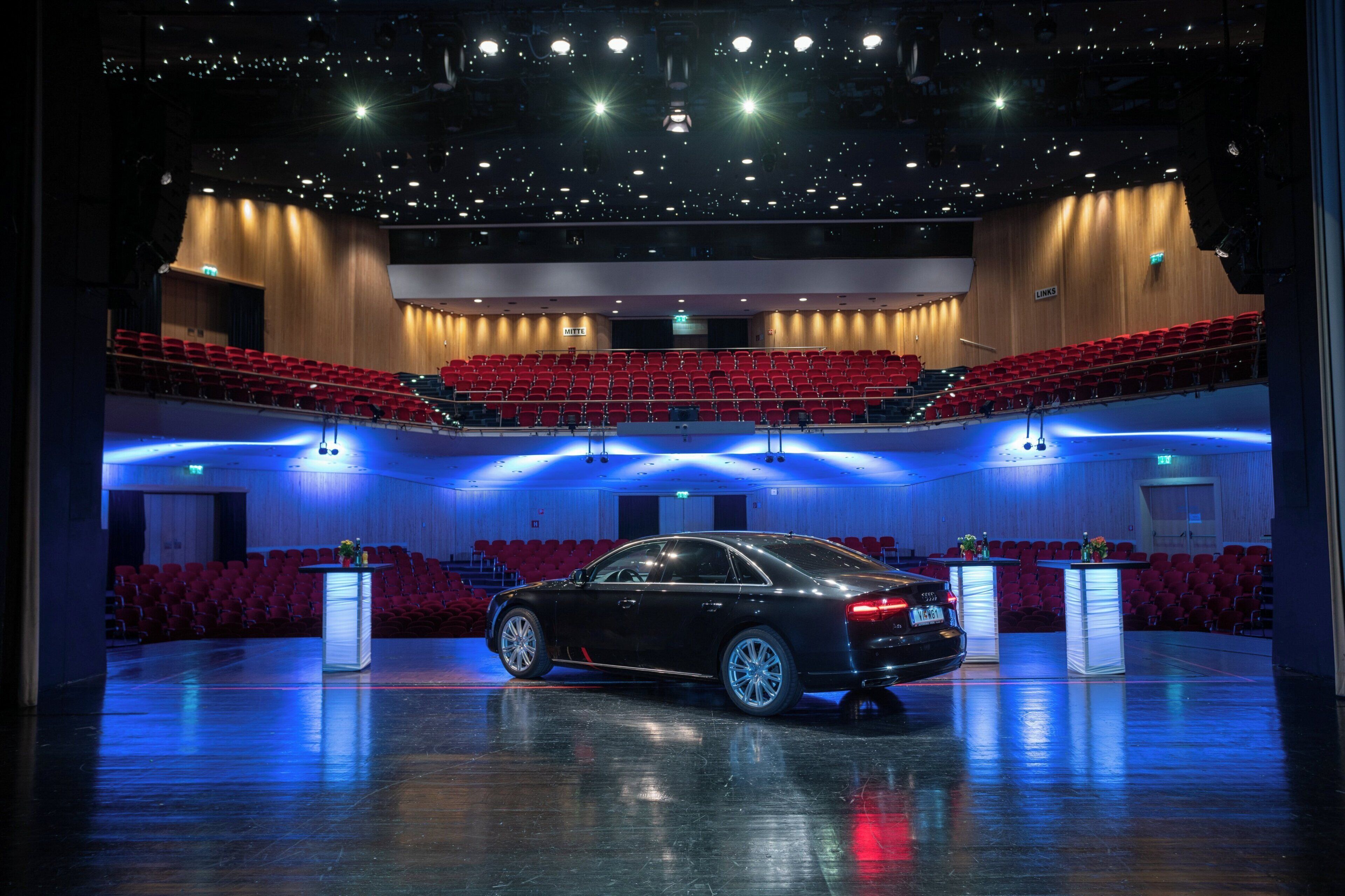 A spacious auditorium with tiered seating, a black luxury car parked in the center, and podiums with microphones.
