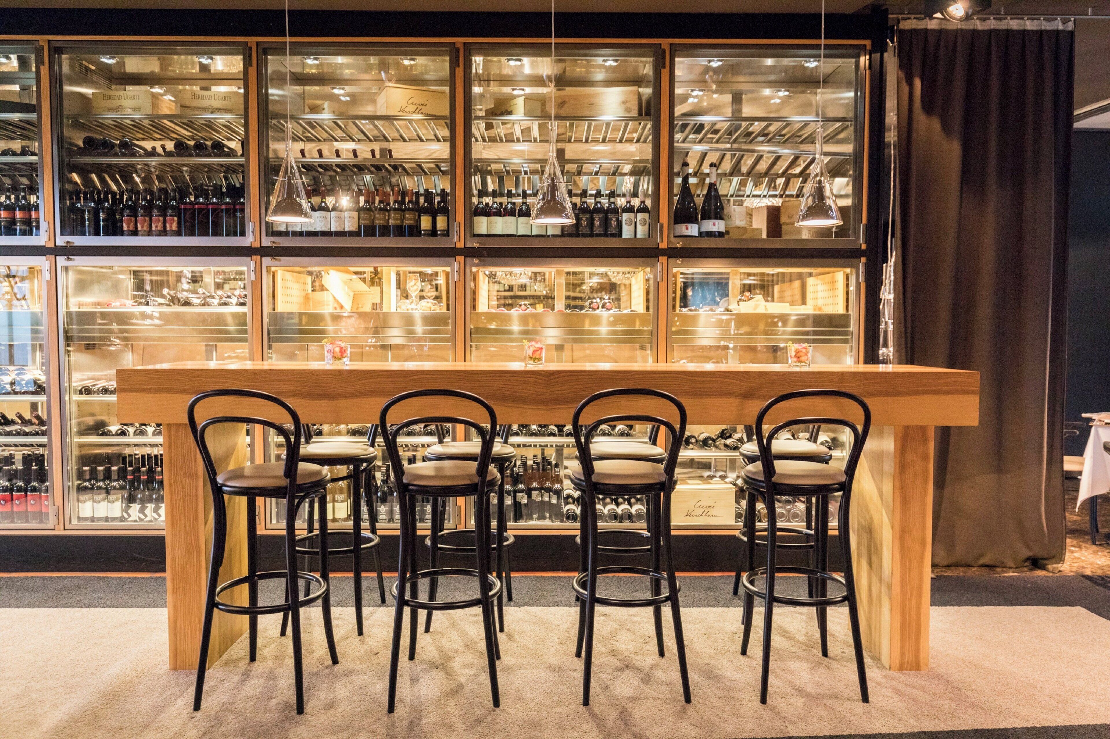 Un espace bar avec un comptoir en bois et quatre chaises noires. Derrière le comptoir, on trouve des étagères remplies de bouteilles.