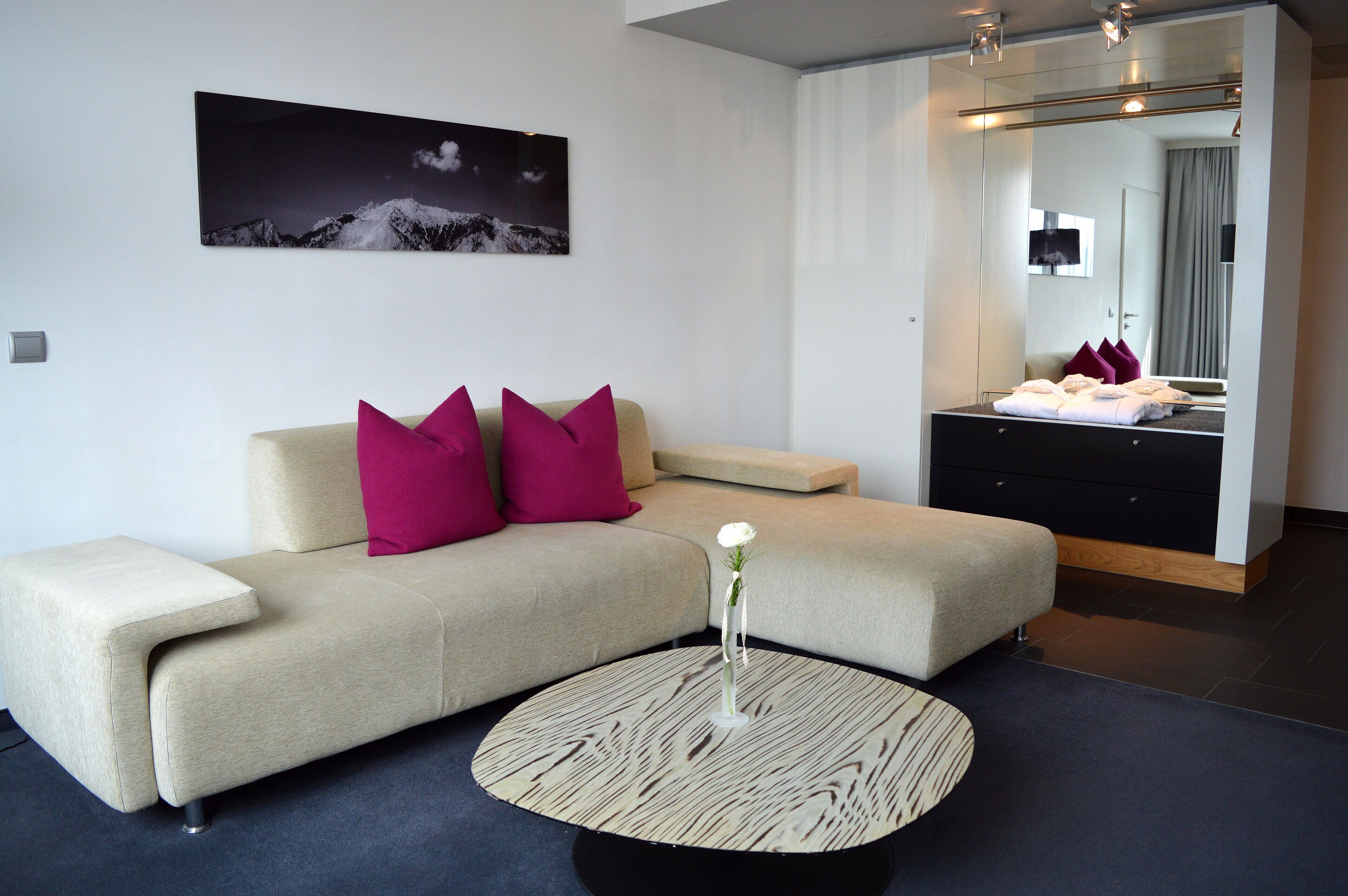 A modern hotel room with a beige sofa, a round coffee table, and a large mirror reflecting a bathroom.