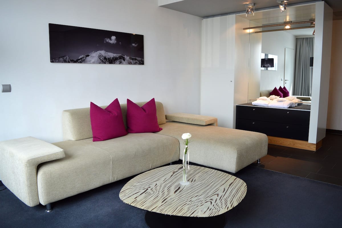 A modern hotel room with a beige sofa, a round coffee table, and a large mirror reflecting a bathroom.