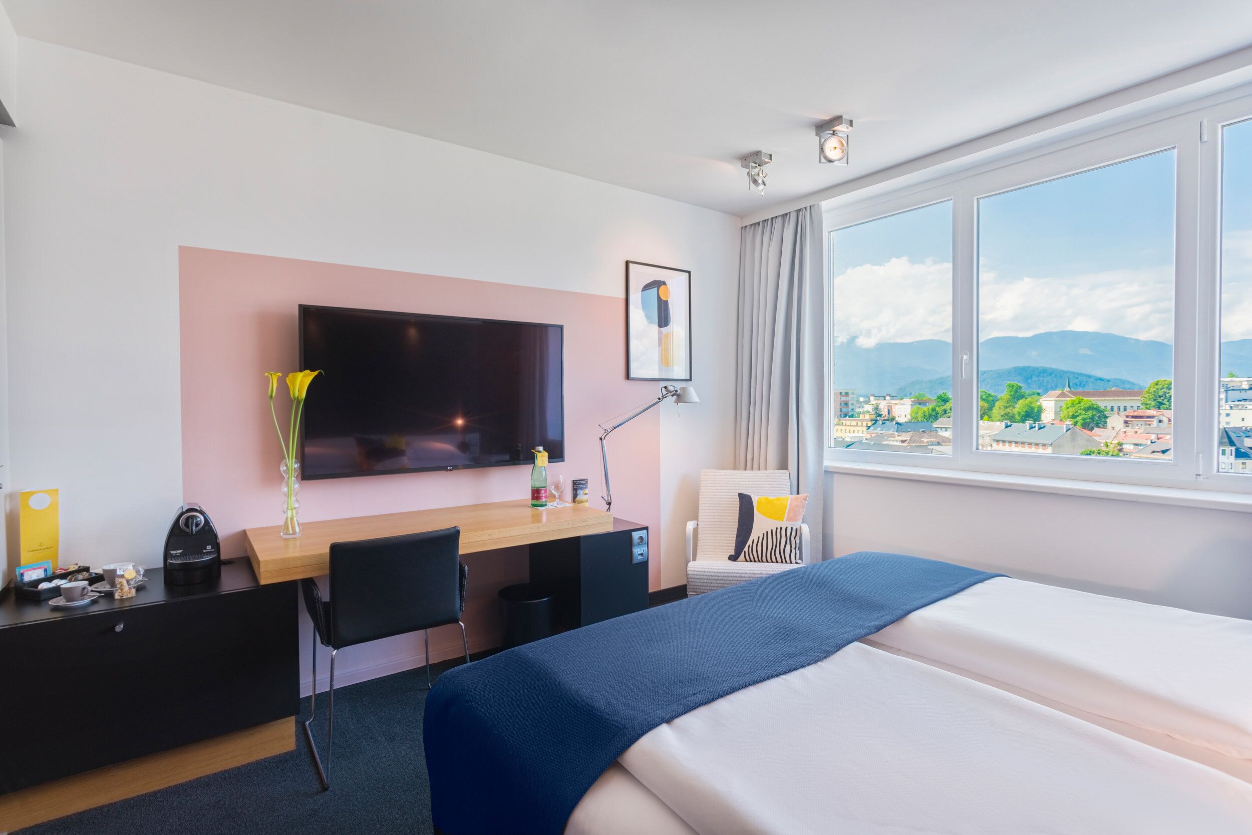 A hotel room with a bed, a desk with a chair, a TV, and a window with a city view.