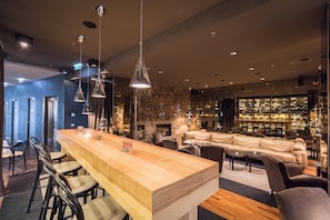 A bar area with a long wooden counter, multiple hanging pendant lights, and a seating area with sofas and chairs.