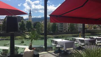Un espacio de estar al aire libre con mesas y sillas, sombrillas rojas, y vistas de un paisaje urbano que incluye una torre de iglesia.