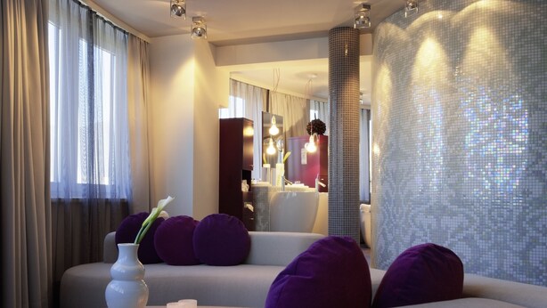 Una habitación de hotel moderna con un sofá, cojines morados, un florero blanco y un espejo.