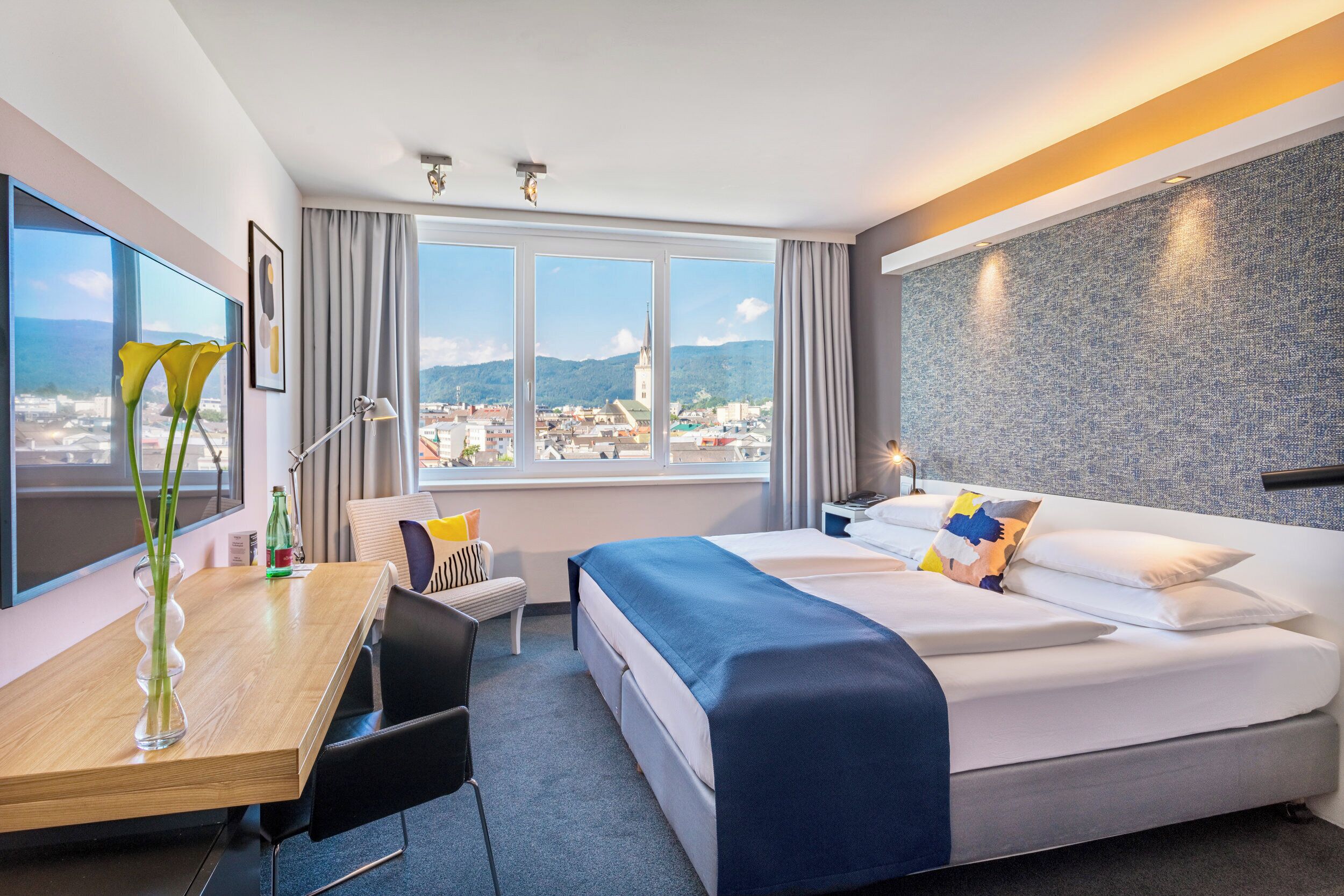 A hotel room with a bed, a desk, a chair, a window with a view of a cityscape, and a wall with a textured pattern.