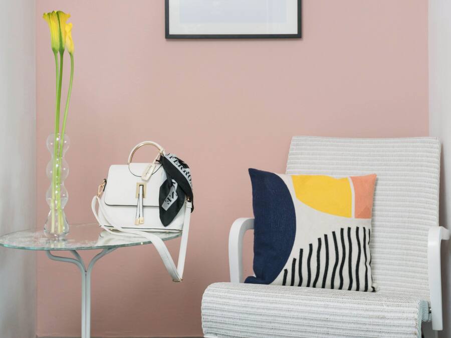 Une pièce avec une table en verre sur laquelle se trouve un vase rempli de fleurs jaunes, un sac à main blanc et une chaise agrémentée d’un coussin à motifs.