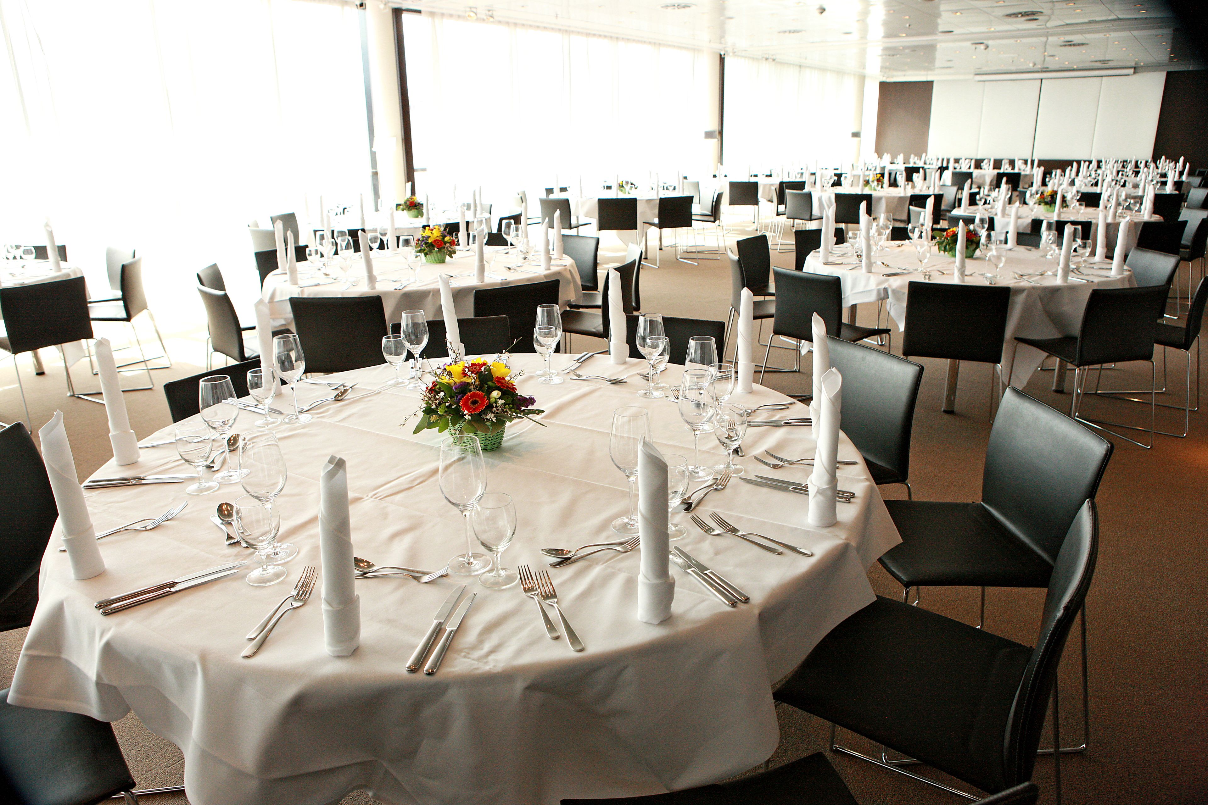 Un salón de banquetes con mesas redondas dispuestas para un evento formal, con manteles blancos, sillas negras y centros de mesa con flores.