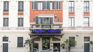 A hotel facade with a sign reading "Hotel Directi," featuring a central entrance with a balcony, two palm trees, and a red brick section above. - Boutique Hotel Dieci (Milan)