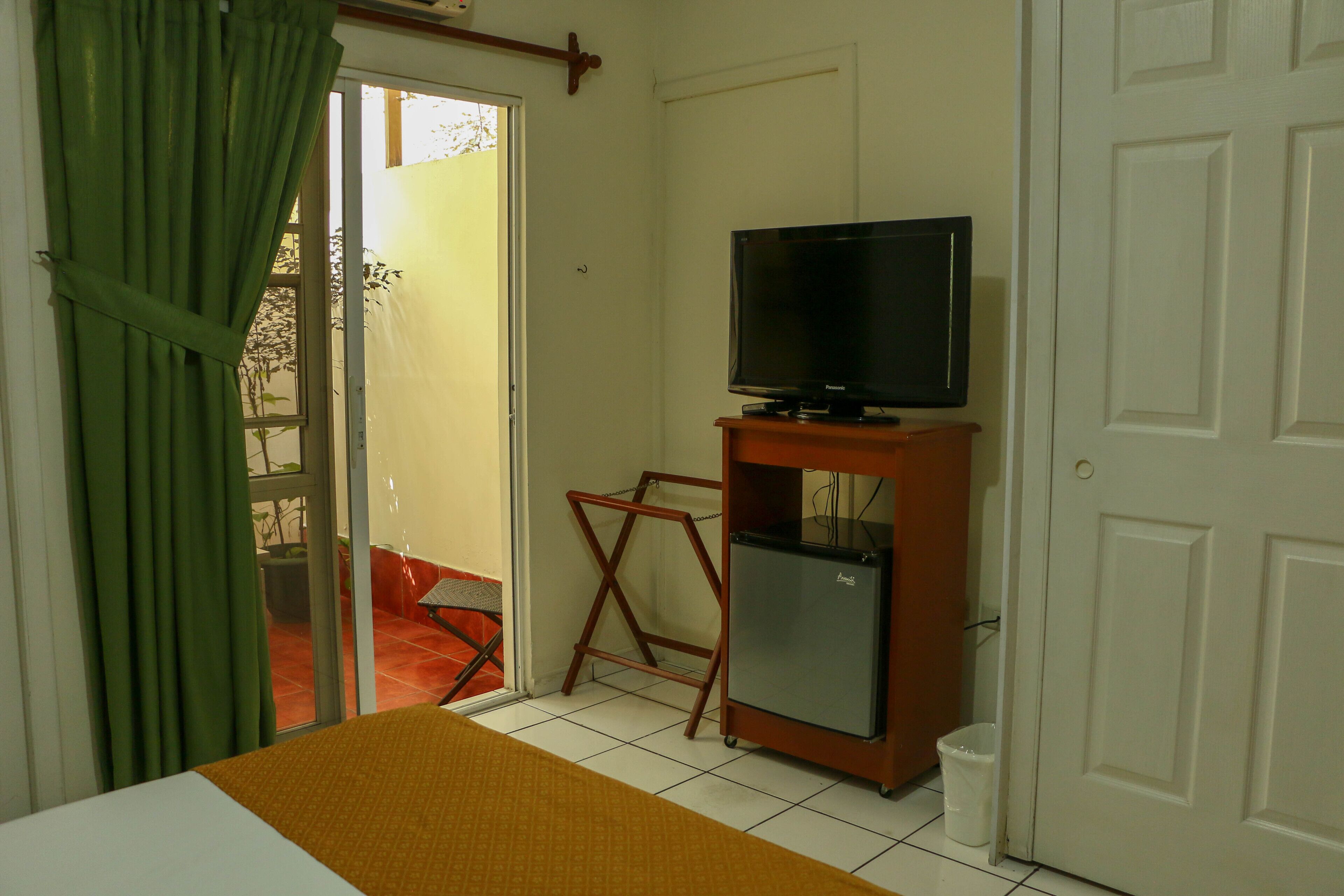 Een kamer met een bed, een televisie op een houten standaard, een kleine koelkast en een deur die uitkomt op een buitenruimte.