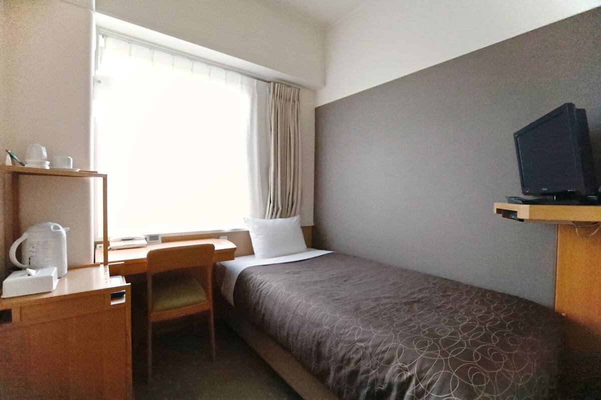 A hotel room with a bed, a desk with a chair, a television on a stand, and a window with curtains.