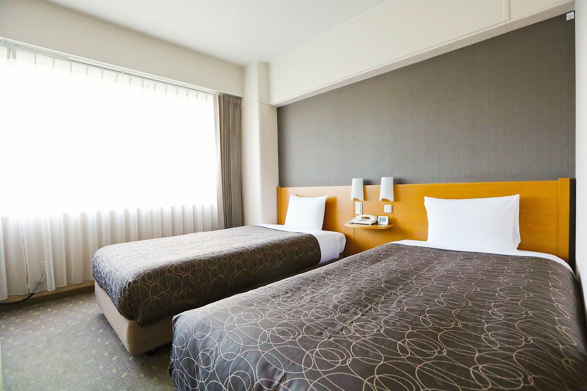 A hotel room with two beds, a large window with curtains, a wooden headboard, and a bedside table with a lamp.