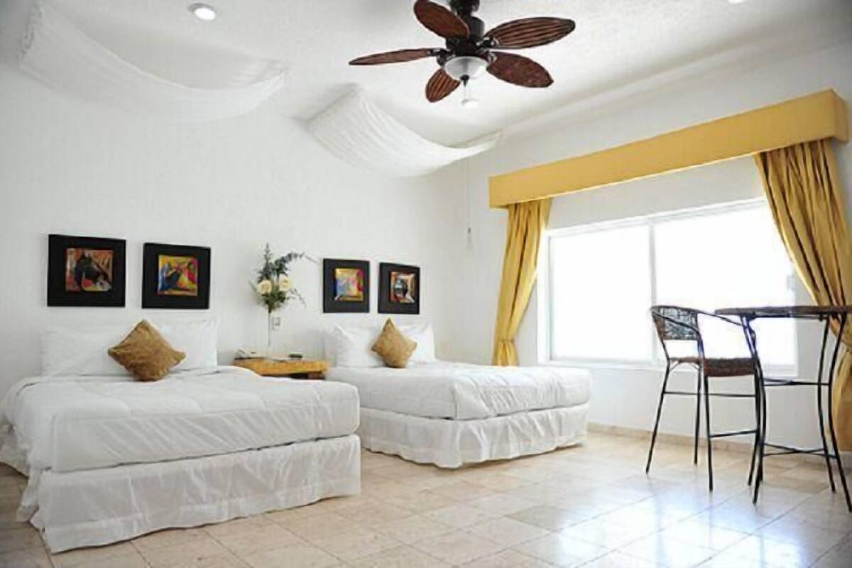 A hotel room with two beds, a ceiling fan, a dining area with chairs, and artwork on the walls.