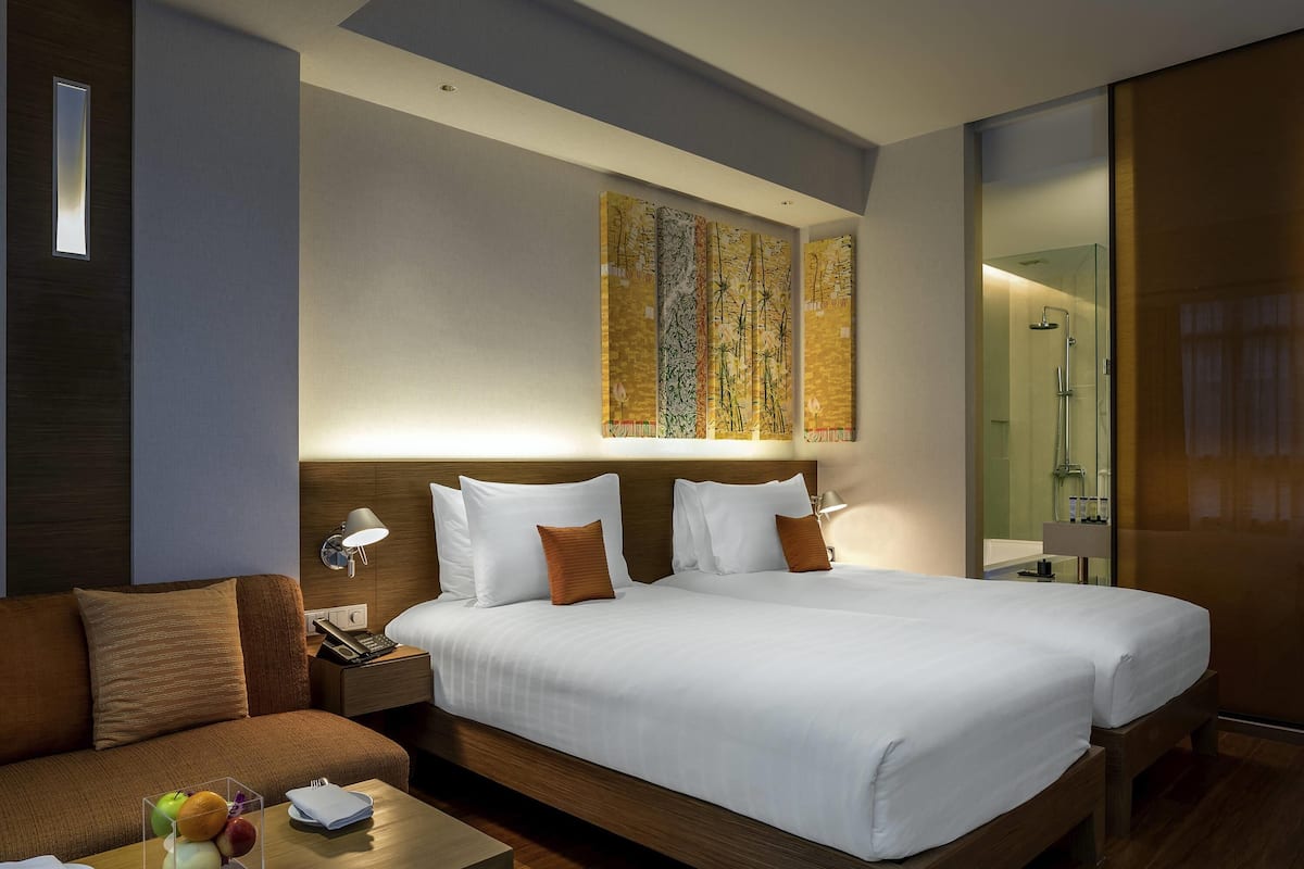 Habitación de hotel con una cama grande, una mesita de noche con lámpara, una pequeña mesa con fruta y un teléfono en la pared.