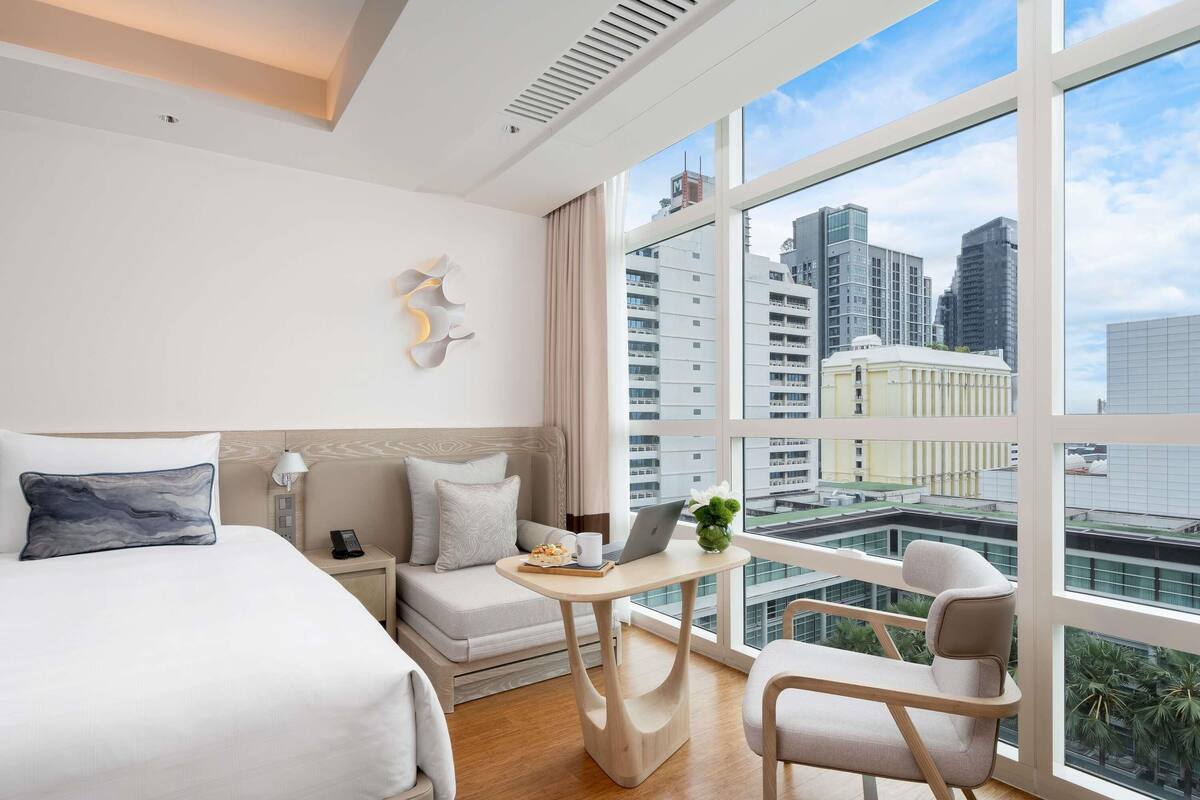 A modern hotel room with a large bed, a small desk, a chair, and a large window offering a city view.
