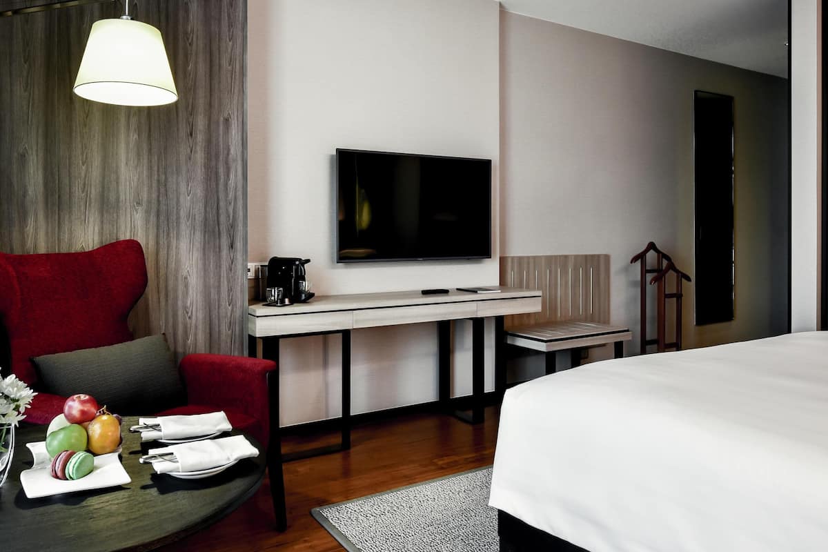 Habitación de hotel moderna con una cama grande, un escritorio con televisor de pantalla plana, un sillón rojo y una cafetera.