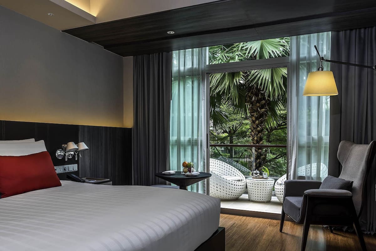 Habitación de hotel moderna con una cama grande, un escritorio con lámpara, una silla y un balcón con vistas a zonas verdes.
