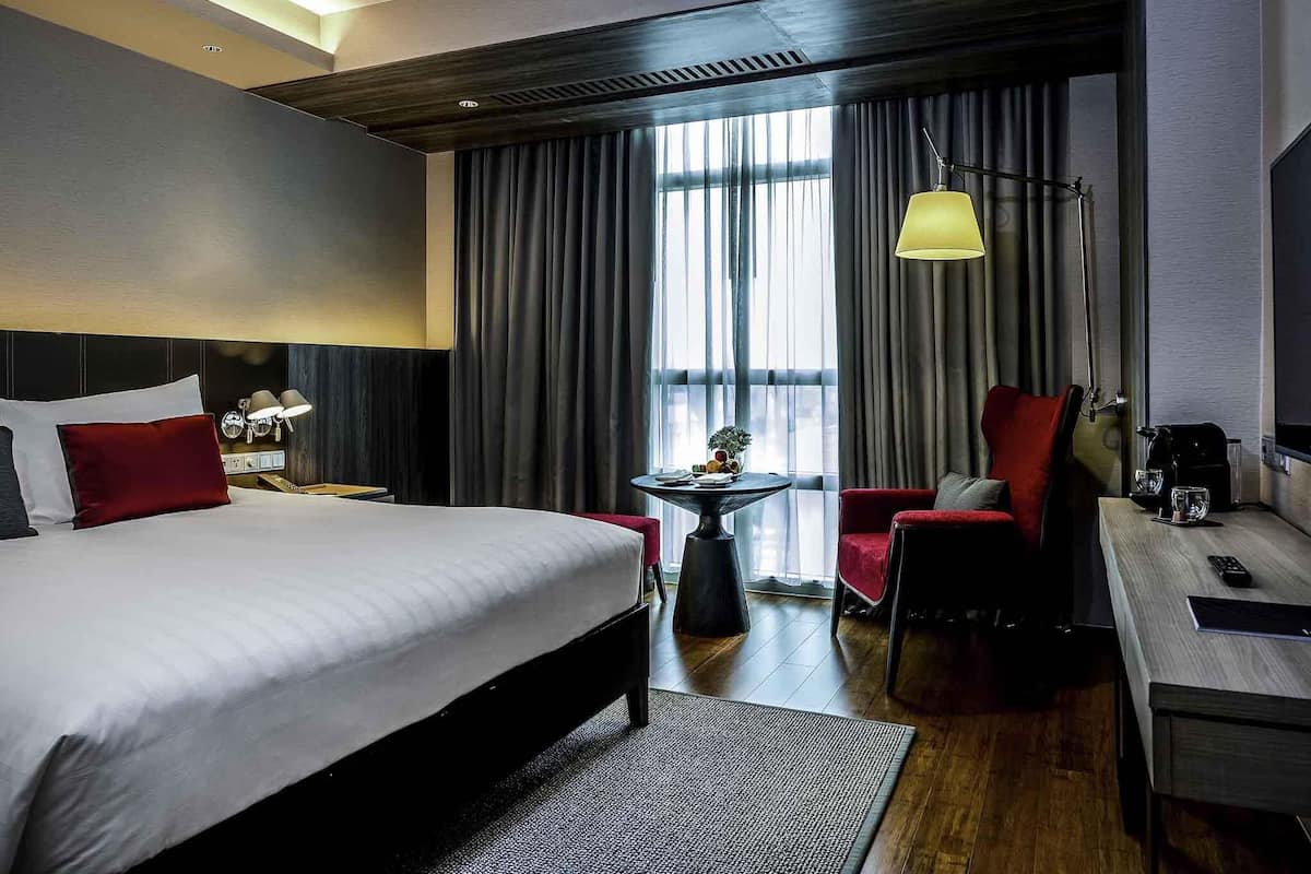 Habitación de hotel con una cama grande, un escritorio, una silla, una mesita de noche con lámpara y vistas a la ciudad a través de amplios ventanales.