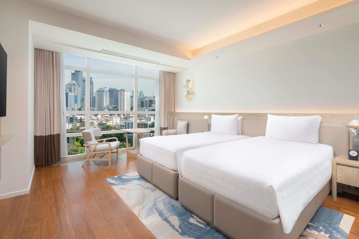 Habitación de hotel con dos camas, un escritorio, una silla, un ventanal grande con vista a la ciudad y un diseño moderno.