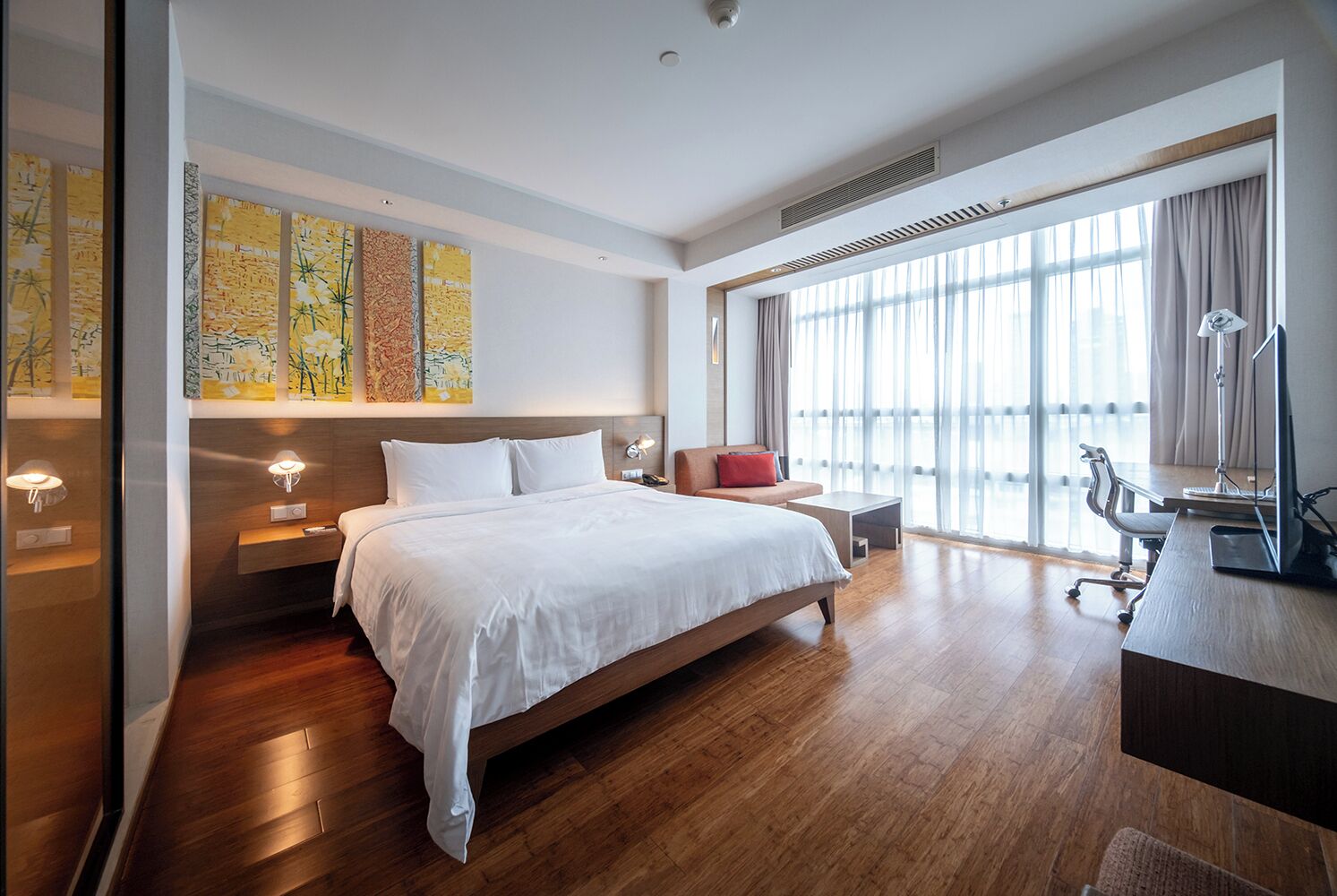 Una camera d'albergo moderna con un letto grande, una scrivania con un computer, una sedia e vista sulla città da ampie finestre.