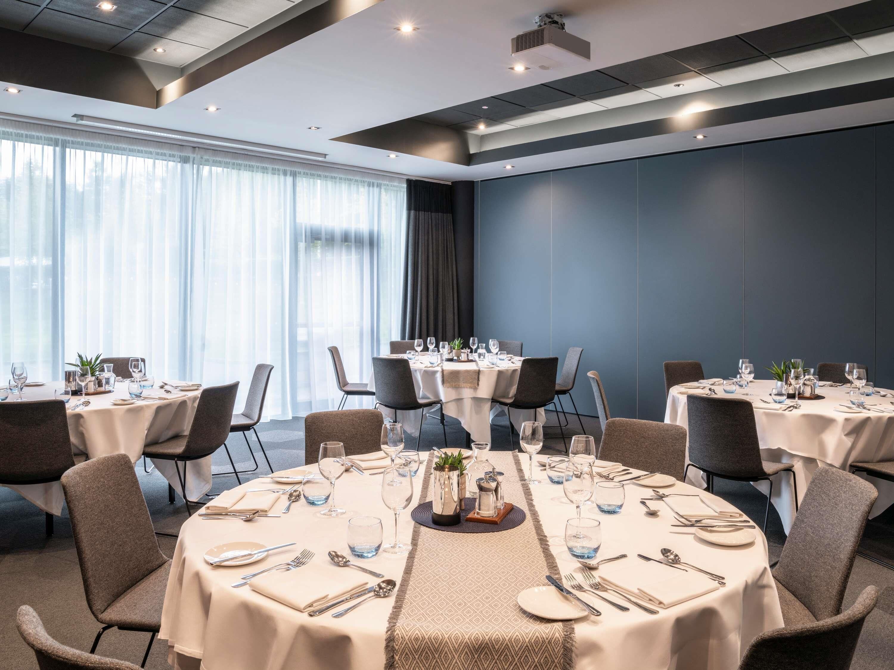 Une salle à manger d’hôtel avec des tables rondes, des chaises et une grande fenêtre agrémentée de rideaux.