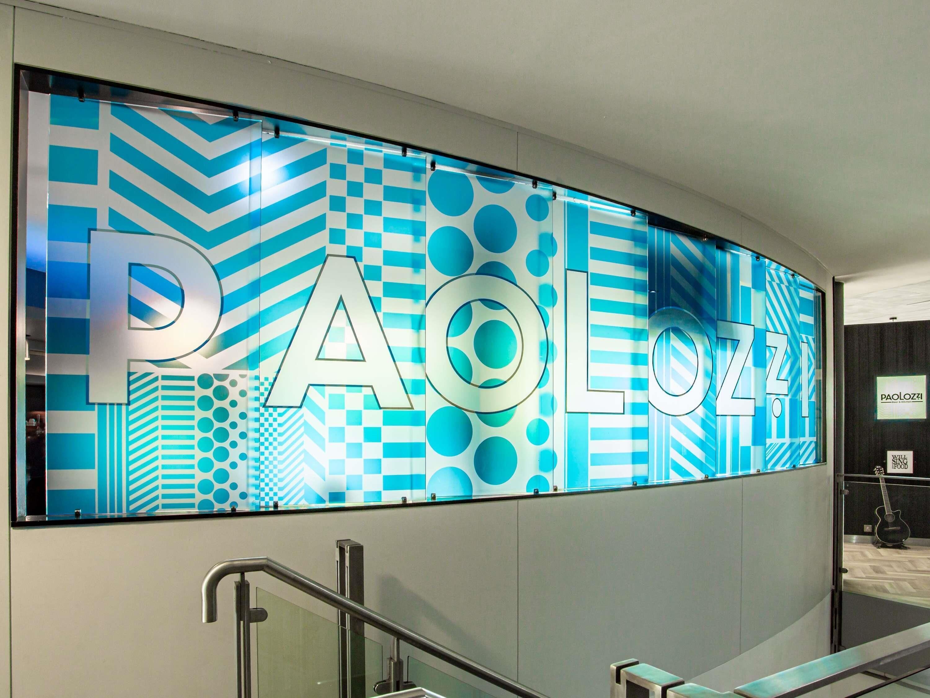 A curved wall with the word 'PAOLOZZI' in large letters, a staircase with metal railings, and a framed picture of a guitar.