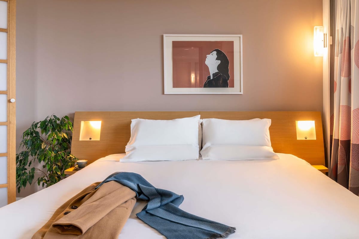 Habitación de hotel con una cama grande, dos lámparas de noche, un cuadro en la pared y una planta al lado izquierdo.