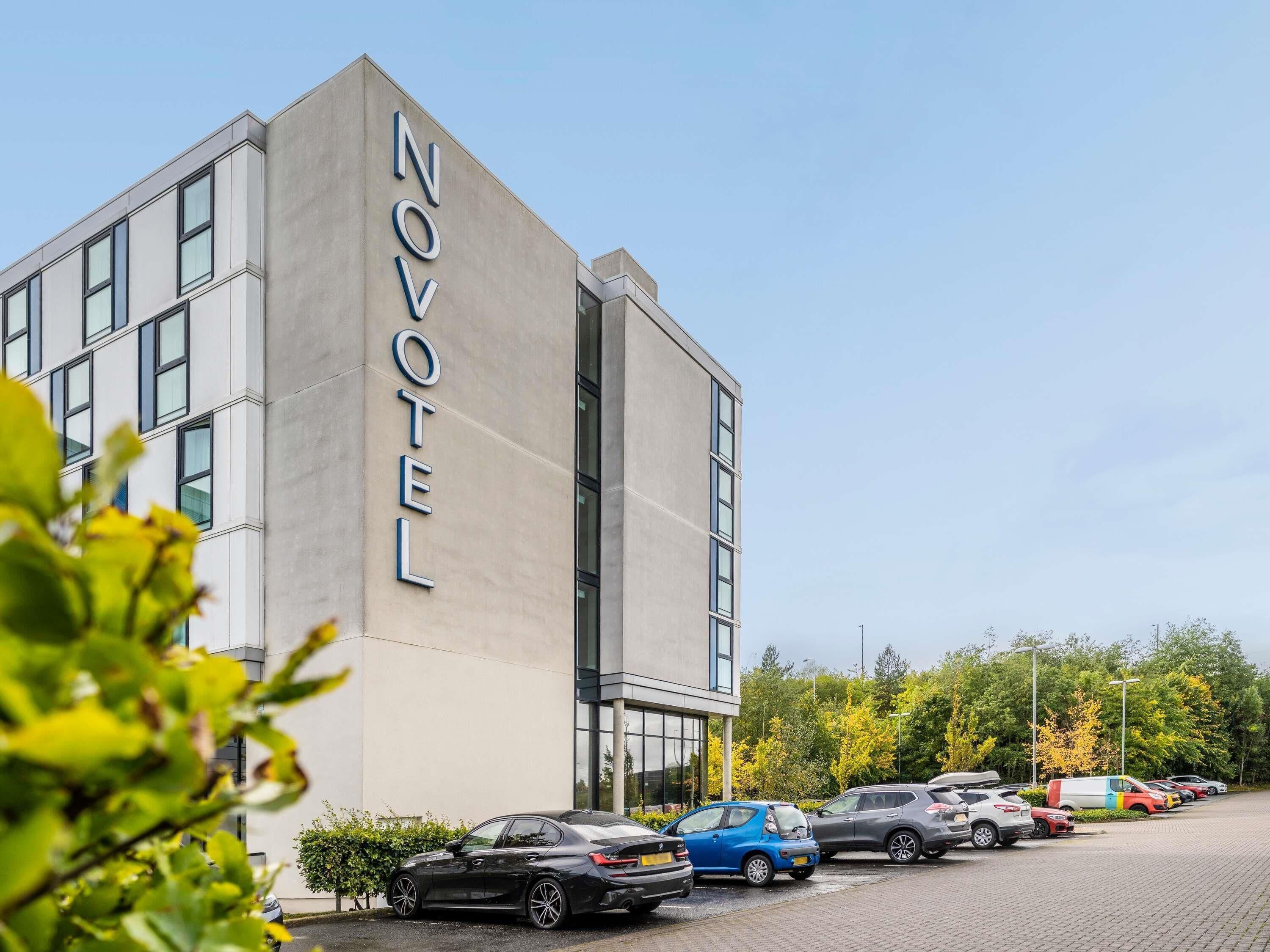 Un moderno edificio de hotel con un llamativo letrero de 'NOVOTEL', un estacionamiento con varios coches y un cielo despejado.