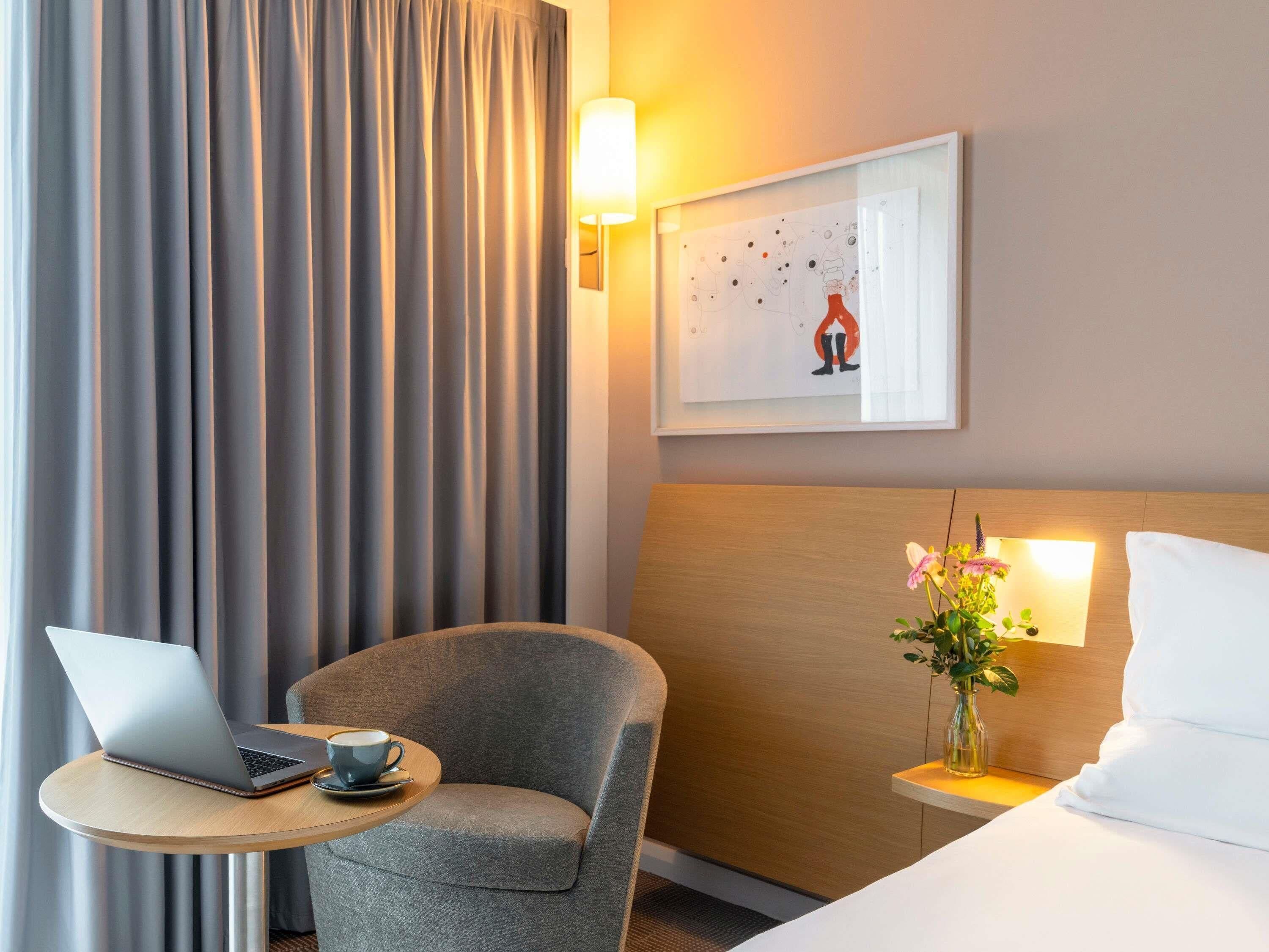 A hotel room with a bed, a desk with a laptop, a chair, a lamp, and a painting on the wall.