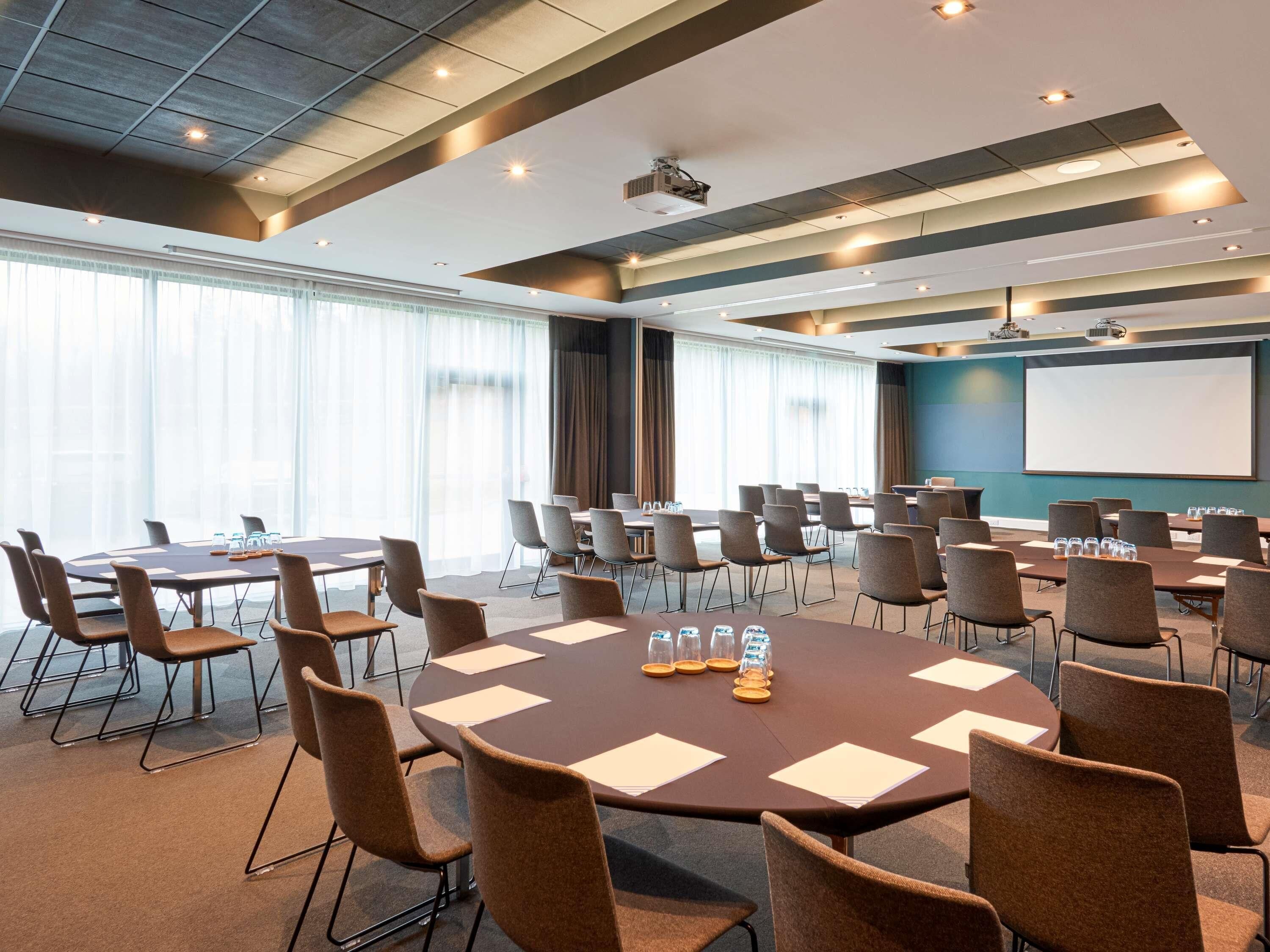 A conference room with multiple tables and chairs, a projector screen, and a large window with curtains.