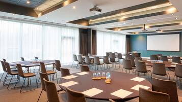 A conference room with multiple tables and chairs, a projector screen, and a large window with curtains.