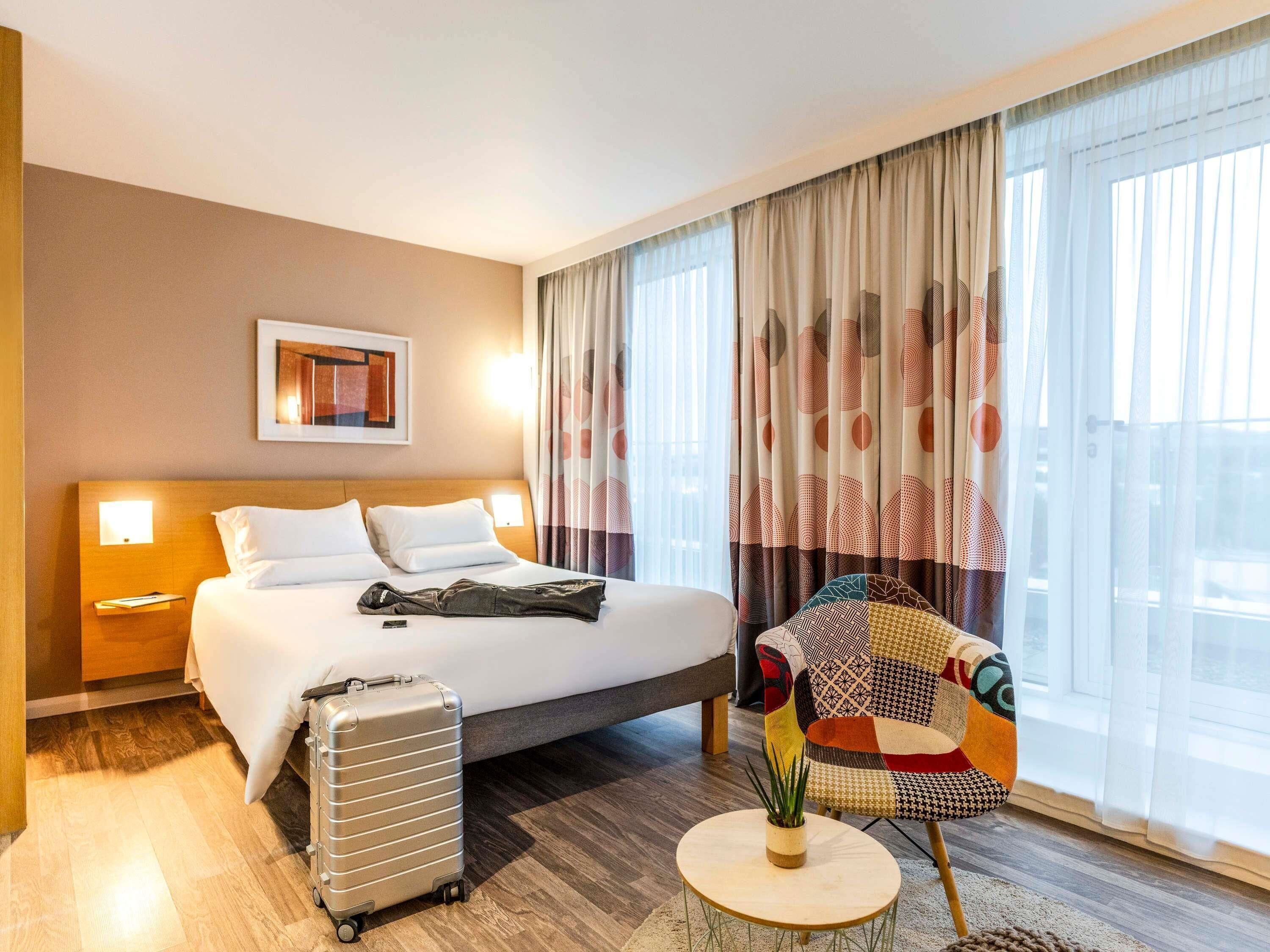 A hotel room with a bed, a suitcase, a chair, and a small table with a plant.