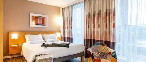 A hotel room with a bed, a suitcase, a chair, and a small table with a plant.